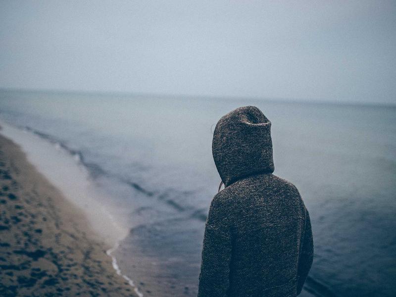 Person in a hooded jacket standing on a sandy beach facing a calm misty ocean